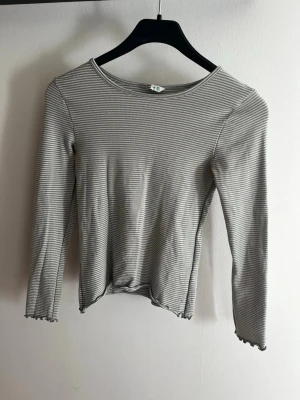 Ljusgrön randig långärmad topp - Snygg långärmad topp i grått och vitt med tunna ränder. Modellen har en rund halsringning och vågiga kanter vid ärmslut och nederkant för en extra detalj. Perfekt att styla med jeans eller kjol. Från cubus.