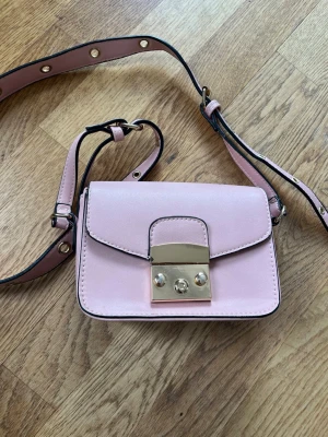 Ljusrosa axelremsväska med gulddetaljer - Söt ljusrosa handväska i slätt konstläder med justerbar axelrem och snygga guldfärgade metalldetaljer på locket. De två inringade gulddetaljerna (bild2) har en liten missfärgning!! Dom information är denna väskan ”låst” innan du öppnar den, för att få upp väskan behöver man trycka dessa två guldknapparna emot varandra!💕