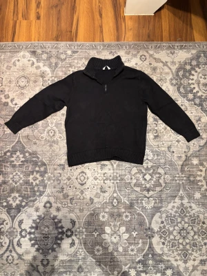 Svart stickad tröja med halv dragkedja - Mysig svart stickad tröja med hög krage och halv dragkedja framtill. Tröjan har långa ärmar och ribbade muddar vid ärmslut och nederkant. Perfekt för lager på lager och en clean look.