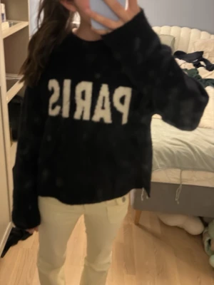 Svart tröja med PARIS-tryck - Cool svart tröja med stort vitt PARIS-tryck på bröstet. Tröjan har en avslappnad passform och långa ärmar, perfekt för en trendig och bekväm look. Materialet ser mjukt och mysigt ut, perfekt för vardagsstilen.