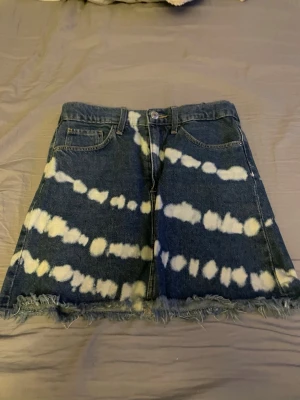 Blå jeanskjol med batikmönster - Cool blå jeanskjol med vitt batikinspirerat mönster och fransig nederkant. Kjolen har klassiska fem fickor och knappstängning framtill. Perfekt för dig som vill sticka ut med en unik stil.