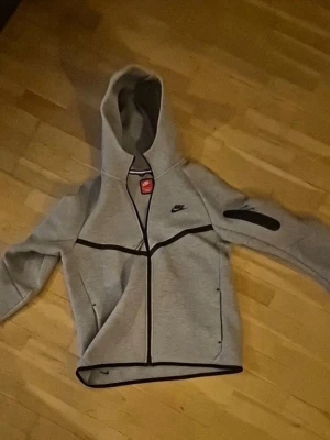 Nike Tech fleece - Snygg grå Nike Tech Fleece hoodie med svarta detaljer och dragkedja framtill. Hoodien har en huva, svarta linjer över bröstet och ärmen samt en praktisk ficka med dragkedja på ena ärmen. Perfekt för en sportig och avslappnad stil.