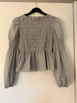 Grå blus med smock och puffärm - Supersöt grå blus med vita små stjärnor som ser ut som prickar, smockad överdel och volang nedtill. Blusen har långa puffärmar med resår vid ärmslut och en rund halsringning. 
