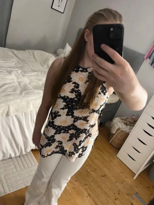 Blommig ärmlös topp - Säljer en blommig ärmlös topp med svart, vit och gul mönstring. Toppen har en rak passform och rund halsringning, perfekt för varma dagar. Materialet känns lätt och luftigt, vilket gör den skön att bära. Säljer även byxorna i bilderna. 