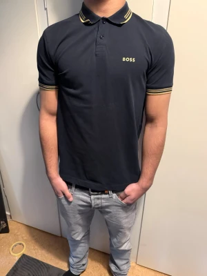 Marinblå BOSS pikétröja med ränder - Snygg marinblå pikétröja från BOSS med gulvita ränder på krage och ärmslut. Klassisk passform med korta ärmar och knappslå framtill. Tillverkad i mjuk bomull som känns skön mot huden. Diskret BOSS-logga på bröstet för en clean look. Nypris: 900. Använd ett par gånger. Storlek S men passar XS.