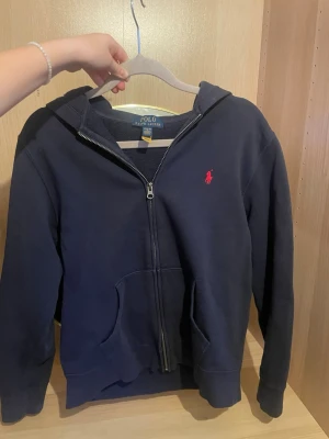 Mörkblå hoodie från Polo Ralph Lauren - Mörkblå hoodie med dragkedja från Polo Ralph Lauren i storlek s.Klassisk design med huva, kängurufickor och rött broderat logotyp på bröstet. Tillverkad i mjuk bomullsblandning som är skön att ha på sig. Perfekt för en avslappnad och stilren look.skriv för fler bilder, pris kan diskuteras!💗