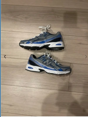 New Balance 530 grå/blå sneakers - Snygga New Balance 530 sneakers i grått och blått med vita detaljer. Skorna har mesh- och syntetmaterial, klassisk N-logga på sidan och vit sula med svarta och blå inslag. Perfekta för en sportig och trendig look.