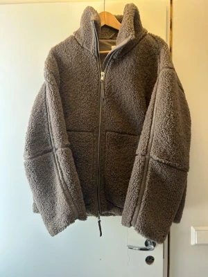 Brun teddyjacka med dragkedja - Mysig brun teddyjacka med hög krage och tvåvägsdragkedja. Jackan har stora fickor framtill och detaljer i matchande brunt tyg längs sömmarna. Perfekt för kalla dagar när du vill vara både varm och stilren.