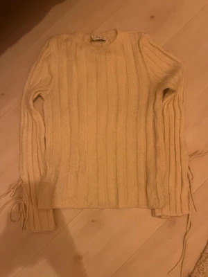 Beige ribbstickad tröja från MNG - Säljer en beige ribbstickad tröja från MNG med rund hals och långa ärmar. Tröjan har dekorativa knytband på sidorna och är gjord i ett mjukt stickat material som passar perfekt för lager på lager. Enkel och stilren design.