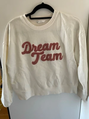 Vit sweatshirt med Dream Team-tryck - Vit sweatshirt från  mango med texten 'Dream Team' i rosa plysch på bröstet. Tröjan har rund halsringning, ribbade muddar vid ärmslut och nederkant samt en avslappnad passform.