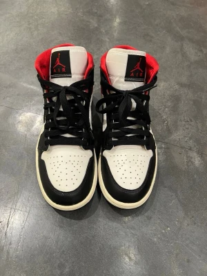 Nike Air Jordan 1 Mid Svart/Vit/Röd - Säljer ett par Nike Air Jordan 1 Mid sneakers i svart, vitt och rött. Klassisk design med svart swoosh, perforerad tåbox och röd vadderad insida. Skorna har svart snörning och ikoniska Air Jordan-loggor på plös och sida. Perfekta för dig som gillar streetwear och retrostil.