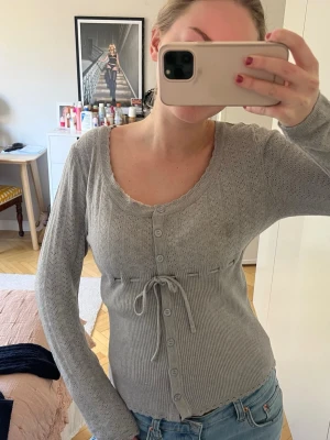 Grå ribbad kofta med knappar - Snygg grå kofta med ribbad nederdel, knappar hela vägen och knytband i midjan. Koftan har lång ärm och rundad urringning, perfekt att slänga över ett linne eller t-shirt. Enkel och stilren design som passar till det mesta.