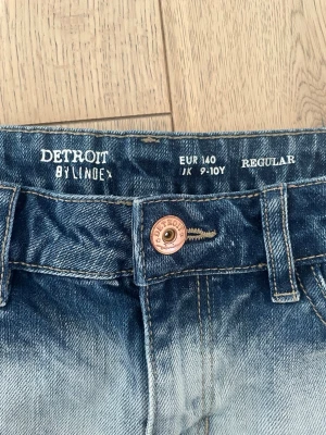 Blå jeansshorts från Detroit by Lindex - Säljer ett par snygga blå jeansshorts från Detroit by Lindex. Shortsen har en cool tvättad look med ljusare partier, råa kanter och klassiska bakfickor. Regular fit och bälteshällor i midjan. Perfekta för sommardagar!