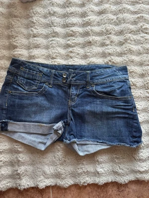 Blå jeansshorts från MNG - Säljer ett par klassiska blå jeansshorts från MNG med råa kanter och femficksmodell. Shortsen har normal passform, knappgylf och detaljerade bakfickor. Perfekta för sommaren och enkla att matcha med olika stilar.