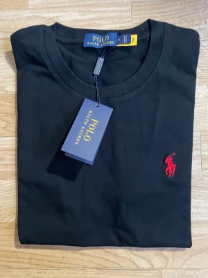 Svart t-shirt från Polo Ralph Lauren - Snygg svart t-shirt från Polo Ralph Lauren med klassisk röd logga broderad på bröstet. T-shirten har rund halsringning och korta ärmar. Tillverkad i mjuk bomull som känns skön mot huden. Perfekt basplagg för en clean och stilren look.