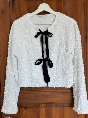 Vit paljettblus med svarta rosetter - Supersnygg vit topp täckt med paljetter och två svarta rosetter framtill. Långärmad modell med rund halsringning och croppad passform. Perfekt för dig som vill sticka ut och addera lite extra bling till din outfit. Använd 1 gång, så jättefint skick 🌸