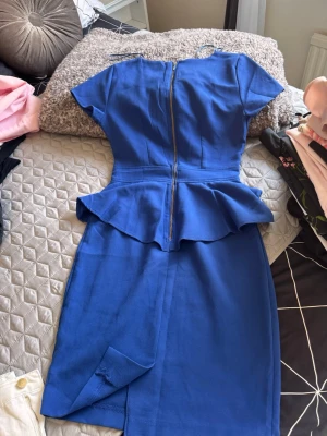 Blå peplumklänning med dragkedja - Snygg blå klänning med peplumdetalj i midjan och korta ärmar. Klänningen har en markerad midja, rak kjol och dragkedja i ryggen. Perfekt för dig som vill ha en stilren och feminin look.