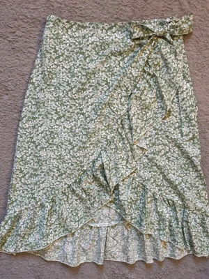 Grön blommig omlottkjol med volang - Söt grön omlottkjol med vitt blommönster och volangkant. Kjolen har en knytning i midjan och är perfekt för vår och sommar. Lätt och luftig modell som ger en romantisk vibe.