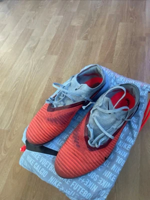 Nike röda och grå fotbollsskor - Säljer ett par Nike phantom 6 elite fotbollsskor i rött och blå med svarta detaljer. Skorna har snörning, låg profil och en modern design med struktur på ovandelen för bättre bollkontroll. Perfekta för dig som vill ha stil och funktion på planen. Fint skick 3100kr nypris så ett kap