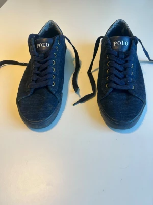 Blå sneakers från Polo Ralph Lauren - Mörkblå sneakers från Polo Ralph Lauren med denimliknande tyg, matchande blå skosnören och metallfärgade öljetter. Klassisk låg modell med Polo-logga på plösen. Perfekta för en avslappnad och stilren look.