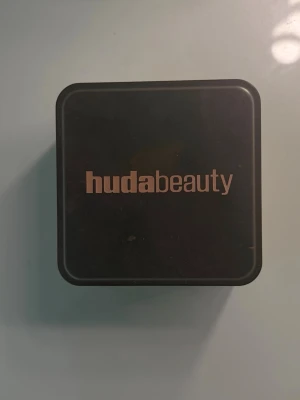 Huda Beauty banana bread puder - Säljer ett löspuder från Huda Beauty i en fyrkantig plastburk med svart lock och transparent underdel. Pudret har en ljus beige ton och ger en matt finish. Perfekt för att fixera din bas och få en jämn look. LITE MER ÄN HÄLFTEN KVAR