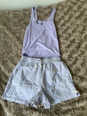 Ljuslila pyjamas-set med linne och shorts - Säljer ett ljuslila pyjamas-set med ett enkelt linne och matchande shorts. Shortsen har vita och ljusgula ränder samt dragsko i midjan för skön passform. Perfekt för chill dagar hemma eller mysiga nätter. Materialet känns mjukt och luftigt.