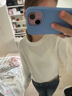 Vit cropped stickad tröja - Säljer en vit cropped stickad tröja med rund hals och långa ärmar. Tröjan har ribbade muddar vid ärmslut och nederkant, vilket ger en snygg och enkel look. Perfekt att matcha med jeans eller kjol för en clean stil.