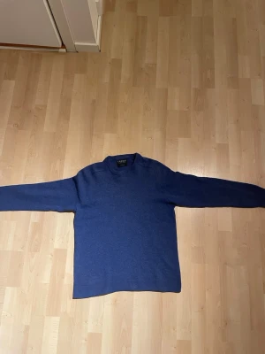 Blå stickad tröja från Canda - Mysig blå stickad tröja från Canda i 100% lammull. Tröjan har rund halsringning och ribbstickade muddar vid ärmslut och nederkant. Perfekt för dig som gillar klassisk och enkel stil. Den har en defekt på ena ärmändsslutet som ni ser på bild. Pris ej hugget i sten. Den är krympt i tvätt så den passar S.