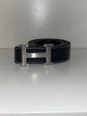 Svart Hermès skärp med H-spänne - Snyggt svart skärp från Hermès med det ikoniska H-spännet i silverfärgad metall. Skärpet är gjort i äkta skinn och har vita kontrastsömmar längs kanterna. Perfekt accessoar för att lyfta vilken outfit som helst.