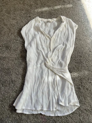 Vit omlottblus i plisserat tyg - Säljer en vit omlottblus med plisserad struktur och knyt i midjan. Blusen har v-ringning och korta ärmar, vilket ger en luftig och avslappnad look. Perfekt till jeans eller kjol för en trendig stil. Har en fläck som säkert går bort med rätt medel