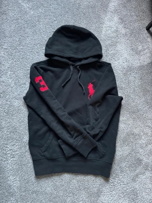 Svart hoodie från Polo Ralph Lauren - Snygg svart hoodie från Polo Ralph Lauren med stor röd logga på bröstet och siffran 3 broderad i rött på ena ärmen. Klassisk känguruficka och justerbar huva med snören. Perfekt för en avslappnad och stilren look.