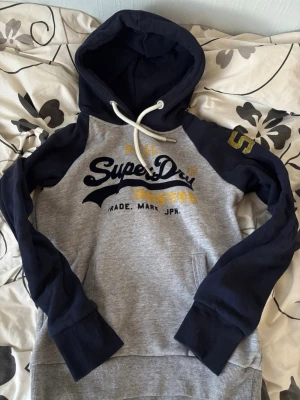 Superdry hoodie size 38 - Snygg hoodie från Superdry i grått med marinblå ärmar och huva. Stor Superdry-logga i mörkblått och gult framtill samt en gul siffra på ärmen. Klassisk känguruficka och vita dragsnören i huvan. Perfekt för en avslappnad streetstyle-look.