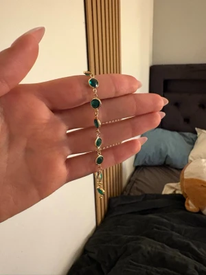 Guldarmband med gröna stenar - Snyggt armband i guldfärgad metall med flera runda, gröna stenar infattade längs hela bandet. Perfekt accessoar för att lyfta din outfit med en färgklick. Lätt och smidigt att bära, passar till många olika stilar.