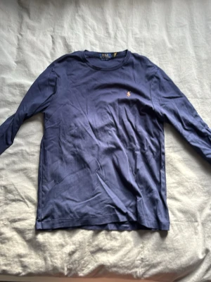 Marinblå långärmad topp från Polo Ralph Lauren - Klassisk marinblå långärmad topp från Polo Ralph Lauren med rund halsringning och den ikoniska broderade loggan på bröstet. Tillverkad i mjuk bomull som känns skön mot huden. Perfekt basplagg till garderoben.