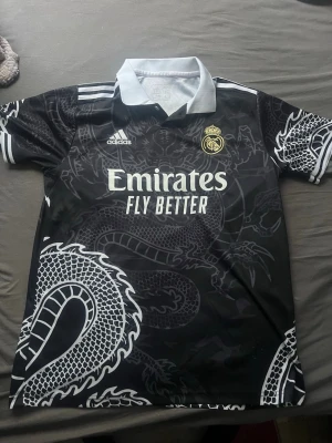 Real Madrid svart draktröja Adidas - Snygg svart Real Madrid matchtröja från Adidas med vit drakdesign över hela tröjan. Tröjan har vit krage, vita detaljer på ärmarna och klubbmärke samt Adidas-logga på bröstet. Tillverkad i lätt och ventilerande material, perfekt för fotboll eller träning.