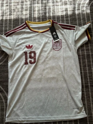 Vit Adidas Spanien matchtröja - Säljer en vit Spanien matchtröja från Adidas med vinröda detaljer, nummer 19 på bröstet och officiellt landslagsemblem. Tröjan har gula och vinröda ränder vid kragen och axlarna samt är tillverkad i ett lätt funktionsmaterial.