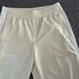 Adidas Adicolor classic Firebird träningsbyxor  - Beiga mjukisbyxor från Adidas med vita klassiska ränder längs benen och broderad logga på sidan. Byxorna har resår i midjan, dragkedjefickor och är gjorda i ett mjukt, stretchigt material som är riktigt skönt. Endast provade