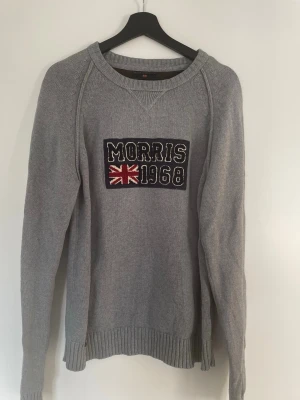 Grå stickad tröja Morris 1968 - Snygg grå stickad tröja från Morris med broderad text 'Morris 1968' och brittisk flagga på bröstet. Tröjan har rund halsringning, långa ärmar och ribbade muddar vid ärmslut och nederkant. Perfekt för dig som gillar klassisk och sportig stil.