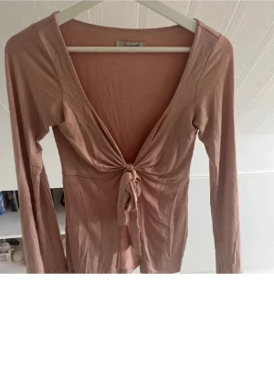 Rosa omlottopp med knyt från Design S - Supersnygg rosa omlottopp från Design S med knyt framtill och djup v-ringning. Toppen har långa ärmar och är gjord i mjuk bomullsmix. Perfekt för dig som gillar en feminin och trendig stil.