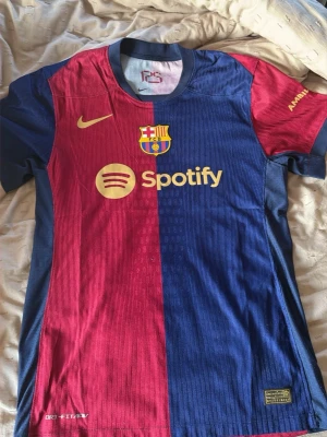 Barcelona matchtröja Nike röd/blå - Säljer en officiell FC Barcelona matchtröja från Nike i rött och blått med klubbmärke, Spotify-logga och gul Nike-swoosh på bröstet. Tröjan har korta ärmar och är tillverkad i ett lätt, ventilerande funktionsmaterial. Perfekt för fotbollsträning eller att bära på läktaren.