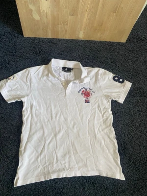 Vit Nautical Center pikétröja - Vit pikétröja från Nautical Center med broderad kompass och flaggor på bröstet samt siffran 8 på ärmen. Klassisk krage och knappar vid halsen. Perfekt för en sportig och avslappnad stil. Hör av er vid frågor eller funderingar 