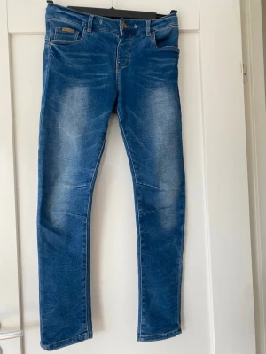 Blå slim fit jeans från LAB Industries, storlek 158 - Snygga blå slim fit jeans från LAB Industries i storlek 158, med klassisk femficksdesign och ljusa slitningar framtill och baktill. Jeansen har normal midja, bälteshällor och knappgylf. Mjukt och stretchigt tyg. Saknas tunt rep, men går att sätta dit eget om så önskas. Jeansen har även resår på insidan för att få till en skön passform. Perfekta för en avslappnad och trendig look.