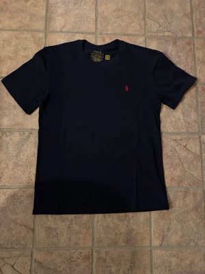 Mörkblå t-shirt från Polo Ralph Lauren - Snygg mörkblå t-shirt från Polo Ralph Lauren med klassisk passform och rund hals. T-shirten har korta ärmar och den ikoniska röda logotypen broderad på bröstet. Perfekt basplagg i mjuk bomull som funkar till allt.