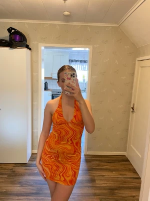 Orange mönstrad halterneckklänning - Säljer en kort, färgstark klänning med halterneck och djup v-ringning. Klänningen har ett psykedeliskt mönster i orange och gult, perfekt för dig som vill sticka ut. Materialet känns lätt och stretchigt, och modellen är tight och smickrande.