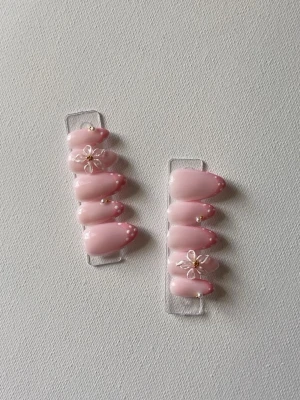 Rosa press-on naglar med blommor - Snygga press-on naglar i ljusrosa med vita prickar och 3D-blommor med gulddetaljer. Naglarna har en oval form och en glansig finish som ger en elegant look. Perfekt för dig som vill ha en söt och trendig nageldesign.