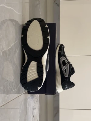 Svarta Dior b30 - Stilrena Dior sneakers i svart med vita detaljer och tjock vit sula. Skorna har en sportig siluett, tydliga kontrastsömmar och stor Dior-logga på sidan. Ovandelen är i textil och lädermix, och sulan är rejält mönstrad för bra grepp. Klassisk snörning och exklusiv känsla.