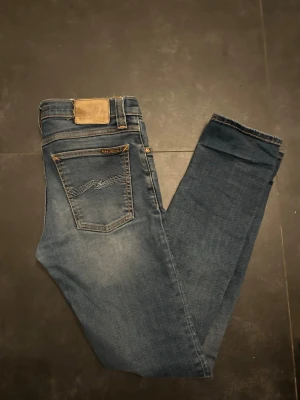 Blå jeans från Nudie Jeans - Säljer ett par klassiska blå jeans från Nudie Jeans med snygga kontrastsömmar och vågig brodyr på bakfickan. Jeansen har normal midja, fem fickor och är gjorda i slitstarkt denim. Perfekta för en avslappnad och trendig look.