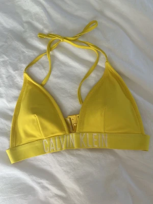 Gul trekants-bh från Calvin Klein - Gul trekantsbikini från Calvin Klein med bred resår och vit logga framtill. Knyt i nacken och spänne ibak. Tillverkad i mjukt syntetmaterial som torkar snabbt och ger en sportig vibe. Pris kan diskuteras.