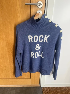 Zadig & Voltaire, Blå stickad tröja med text och knappar - Superfin blå stickad tröja med vit text 'ROCK & ROLL' framtill, från Zadig & Voltaire, aldrig använd , helt ny! Tröjan har hög krage och dekorativa vita knappar längs ena axeln. Långa ärmar och ribbade muddar vid ärmslut och nederkant. Perfekt för dig som gillar en cool och avslappnad stil. Nypris 2500kr.