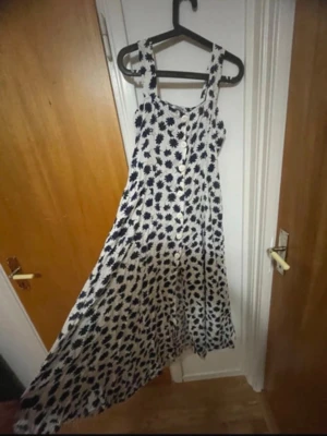 Vit långklänning med blått blommönster - Långklänning i vit med blått blommönster och breda axelband. Klänningen har knappar framtill och ett luftigt, svepande tyg som passar perfekt för varma dagar. Snygg och flowy siluett som ger en avslappnad men ändå chic vibe.